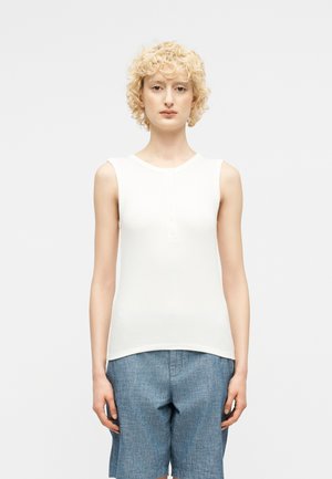 Άτομο με ξανθά σγουρά μαλλιά, ντυμένο με λευκό αμάνικο τοπ και μπλε σορτς μέχρι το γόνατο, στέκεται μπροστά από ένα απλό φωτεινό φόντο.