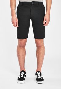 Shorts neri di cotone con taglio dritto, passanti per cintura e tasche frontali. Abbinati a scarpe da ginnastica bianche e nere con allacciatura.
