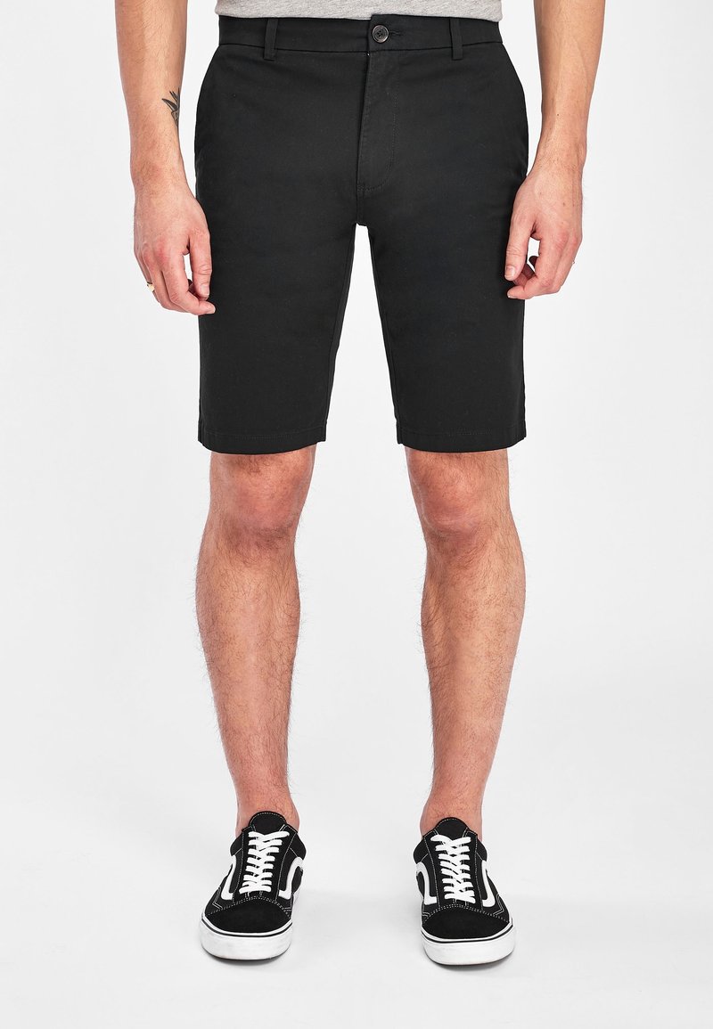 Shorts neri di cotone con taglio dritto, passanti per cintura e tasche frontali. Abbinati a scarpe da ginnastica bianche e nere con allacciatura.