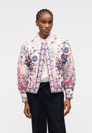 Donna con capelli intrecciati che indossa una giacca trapuntata bianca con motivo floreale e dettagli rosa, blu e marroni sopra una maglietta bianca e pantaloni neri.