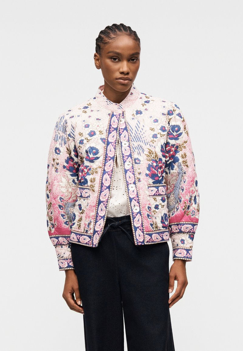 Femme aux cheveux tressés portant une veste matelassée blanche à motif floral avec des accents roses, bleus et marron, par-dessus un top blanc et un pantalon noir.