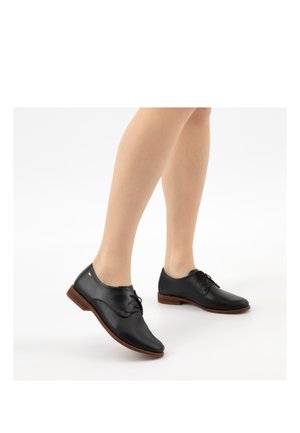 Gambe che indossano scarpe oxford in pelle nera traforata con suole marroni, su sfondo bianco, un piede leggermente sollevato.