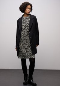 Cappotto nero su un vestito grigio con stampa leopardata, abbinato a collant neri e stivaletti alla caviglia. Tessuto liscio con una trama testurizzata e bottoni.