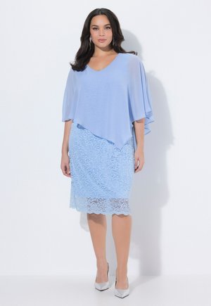 Frau trägt einen hellblauen Spitzenrock und ein passendes fließendes Oberteil, steht vor einem schlichten weißen Hintergrund mit silbernen Absätzen.