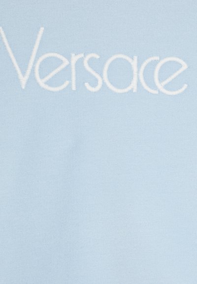 Ανοιχτό μπλε ύφασμα με λείο υφή που διαθέτει λευκό κεντημένο κείμενο που γράφει "Versace." Επικεντρώνεται σε μίνιμαλ σχέδιο και καθαρή επωνυμία.