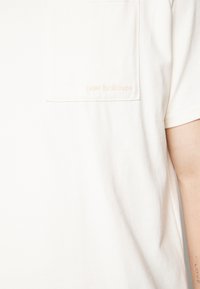 Krémové bavlněné tričko s přední kapsou a vyraženým logem "new balance" ve stejné barvě. Krátké rukávy, jednoduchý design.