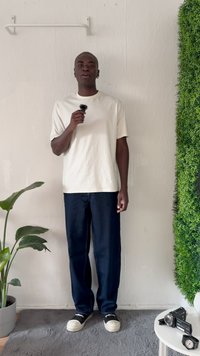 Vit bomull t-shirt, marinblå denimbyxor, svarta slip-on skor. Inomhusmiljö med grönska och en vägghylla i bakgrunden.