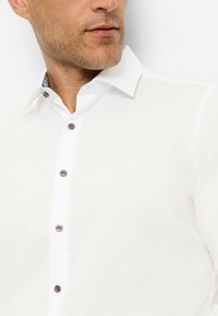 Weißes Hemd aus glattem Stoff mit spitzem Kragen und einer Knopfleiste mit dunklen Knöpfen, akzentuiert durch einen gemusterten Inneng kragen.