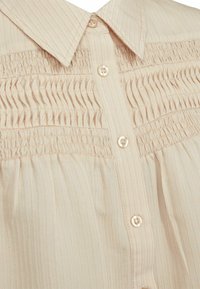 Heledbeez värvi särk ribakanga tekstuuriga, mis on varustatud terava kraega, smockimise detailiga ja väikeste nuppudega ees.