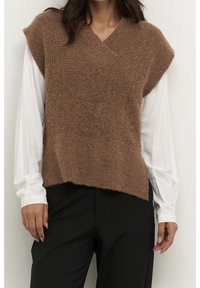 Vrouw draagt een bruin gebreid mouwloos vest over een witte blouse met lange mouwen en zwarte broek, staand tegen een lichte muur.