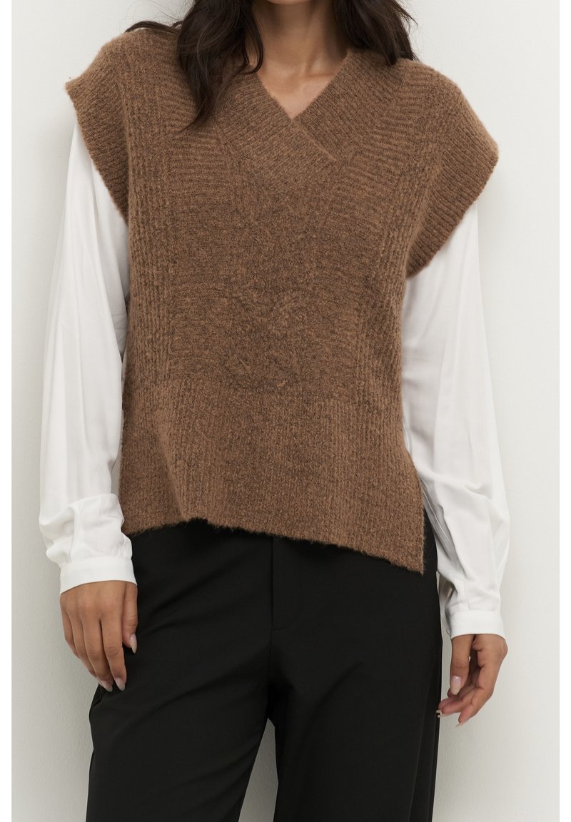 Vrouw draagt een bruin gebreid mouwloos vest over een witte blouse met lange mouwen en zwarte broek, staand tegen een lichte muur.