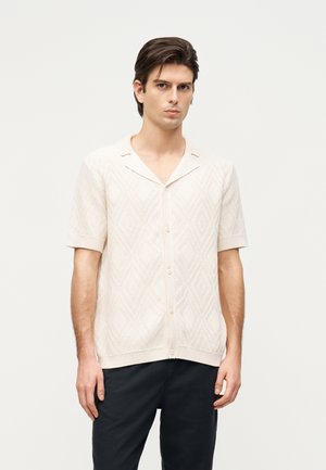 Jonge man draagt een kortärmelig, crèmekleurig, gestructureerd overhemd met knopen en een inkepingkraag, en donkere broek, staand tegen een effen achtergrond.