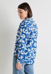 Camisa azul de botones con patrones de hojas blancas, de mangas largas y con dobladillo curvado. Fabricada con una tela ligera, adecuada para el uso casual.