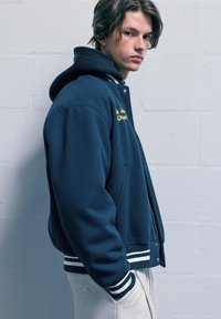 Námorně modrá bomber bunda s bílými pruhovanými manžetami, s vyšívanou nášivkou. Měkká tkanina a kapuce pro teplo a pohodlí.