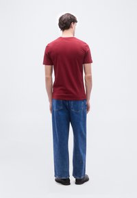 Άνδρας που φοράει κόκκινο κοντομάνικο πουκάμισο, μπλε τζιν και μαύρα παπούτσια, στέκεται με την πλάτη προς το μέρος μας μπροστά σε απλό ανοιχτό φόντο.