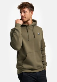 Sudadera verde oliva de tejido suave, con un bolsillo frontal, cordones y un pequeño logo en el pecho. Diseño simple y casual.