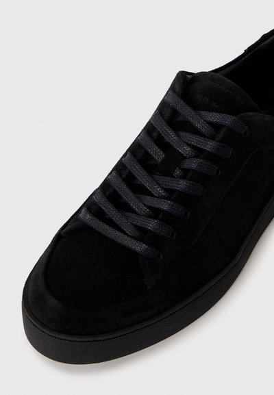 Sort ruskinds sneakers med struktureret overflade, flade sorte snørebånd og en glat gummisål. Minimalistisk design uden synlig branding.