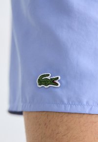Lyseblå shorts laget av glatt stoff, med et grønt krokodillelogo-patch med hvit kant på venstre søm.