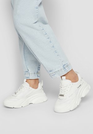 Ben iført lyseblå jeans og hvide chunky sneakers med "Steve Madden"-mærke, stående på en ensfarvet lysegrå baggrund.