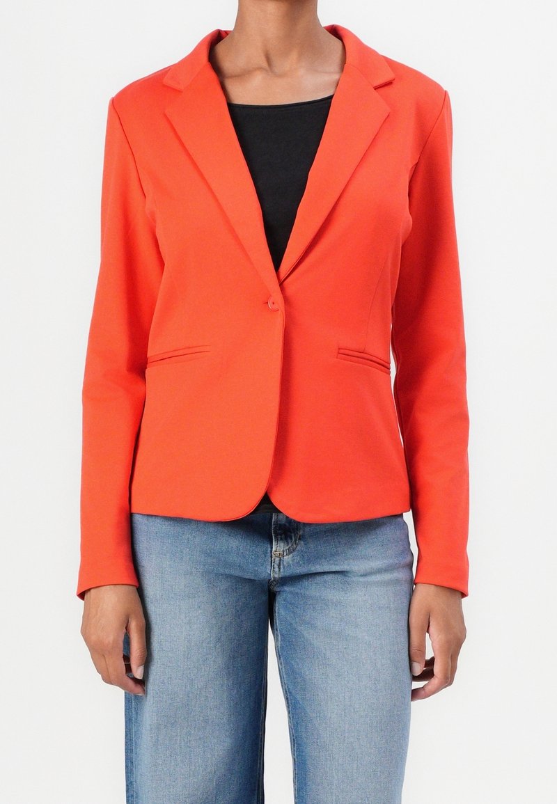 Blazer naranja con cierre de un solo botón, solapas con muesca, dos bolsillos frontales, diseño estructurado y suave textura de tela. Llevado sobre una blusa negra.