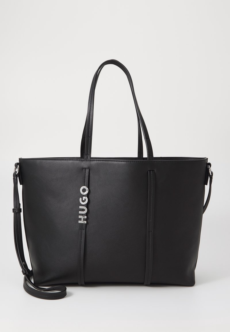 Sac cabas en cuir noir avec deux poignées, bandoulière ajustable et breloque logo "HUGO" argentée sur le devant.