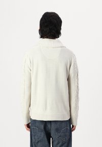 Krämfärgad stickad tröja med hög krage, med kabelstickning på ärmarna och ribbade muddar.
