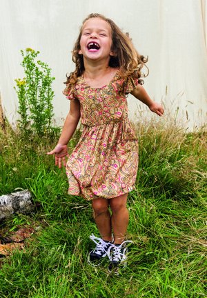 Meisje in een bloemenprint, rozerood en gouden paisleypak met gerimpelde mouwen, lachend terwijl ze zwarte sneakers met witte veters draagt, staande in het gras.