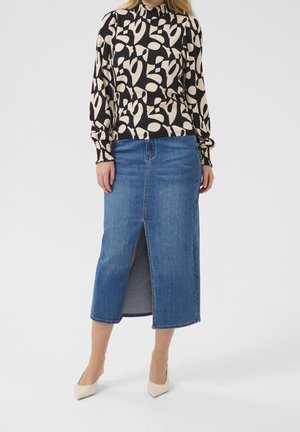 Vrouw draagt een zwarte en beige top met abstracte print, lange mouwen en een hoge hals, gecombineerd met een blauwe denim midi-rok met voorste split en beige hakken.