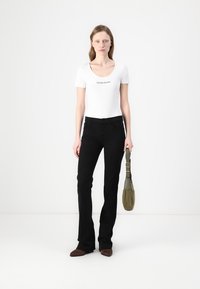 Valge sobiv t-särk, mille rinnal on trükitud "GUESS JEANS", kombineeritud mustade flared teksade ja väikese oliivrohelise käekotiga.