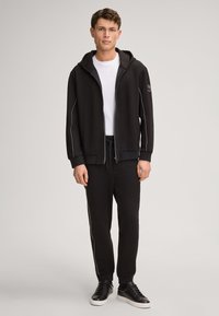 Zwarte sweatshirt met capuchon, ritssluiting en witte accenten. Gecombineerd met bijpassende joggingbroek en zwarte sneakers, met een relaxte pasvorm.