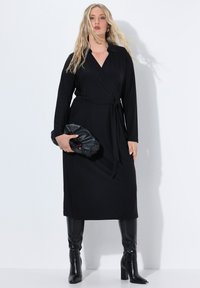 Zwarte wikkeldress van zachte stof, met lange mouwen, een kraag en een tailleband. Gecombineerd met zwart laarzen tot de knie en een zwarte clutch.