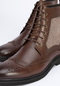 Botas de cuero marrón hasta el tobillo con detalles de brogue y paneles de tela de espiga. Presentan una punta redondeada y un diseño de cordones con ojales.