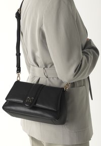 Borsa a tracolla in pelle nera con patta ripiegata e dettagli in oro, caratterizzata da una lettera "M". Cinghia attaccata per il trasporto a spalla.
