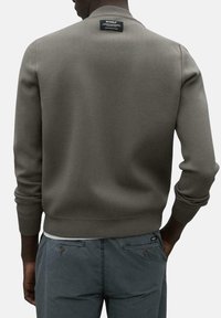 Suéter gris de punto con cuello alto, puños y dobladillo acanalados, que presenta una etiqueta negra en la parte posterior. Lleva puesto sobre pantalones oscuros.