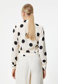 Blusa a maniche lunghe in seta color crema, con grandi pois neri, una vestibilità aderente e un dettaglio in vita annodato. Texture morbida, polsini con bottoni.