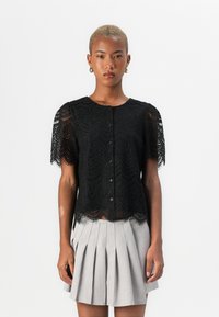 Blusa nera in pizzo con maniche corte, caratterizzata da un orlo scallop e chiusura frontale a bottoni, sovrapposta a una gonna grigia plissettata.