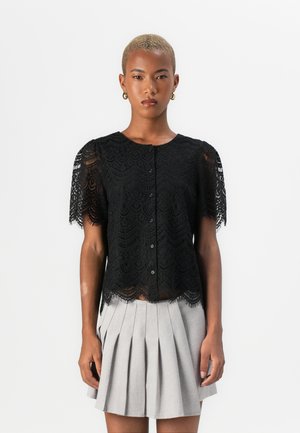 Black lace blouse with short sleeves, featuring a scalloped hem and button front closure, layered over a pleated grey skirt.