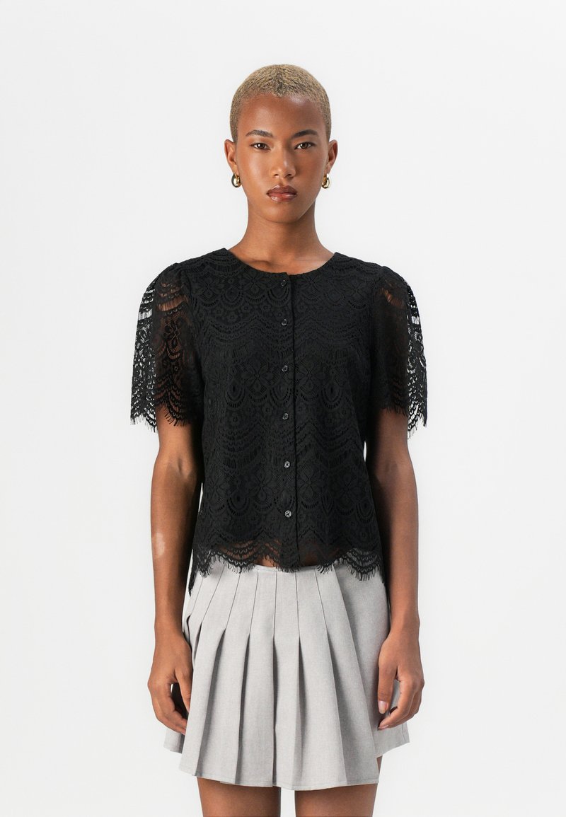 Blusa nera in pizzo con maniche corte, caratterizzata da un orlo scallop e chiusura frontale a bottoni, sovrapposta a una gonna grigia plissettata.
