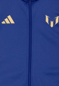 Blauwe ritsjack gemaakt van gladde stof met gouden accenten, waaronder het Adidas-logo en een opvallend embleem op de borst.