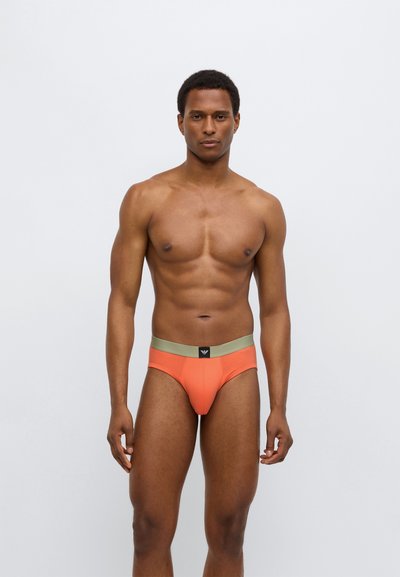 Herrkalsonger i livfull orange tyg med ett mjukt grönt midjeband och en svart loggdetalj. Figursydd design, slät textur, minimal täckning.