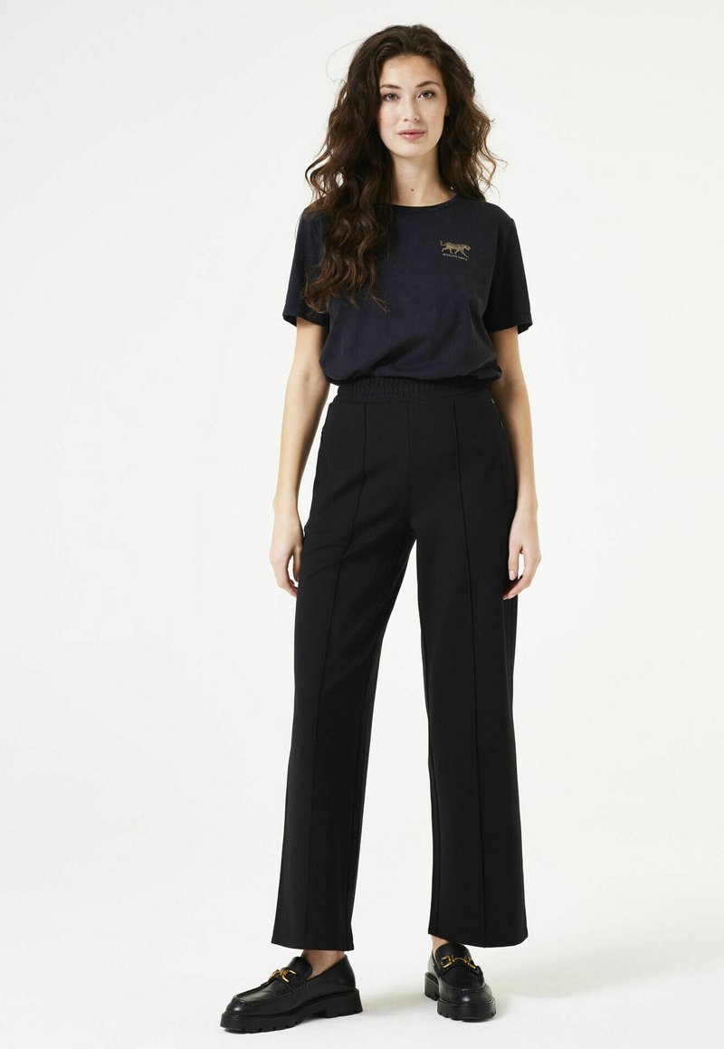 Zwarte wijde broek met een gladde textuur, voorzien van voorste naadlijnen en een elastische tailleband. Gekleed met een marineblauw T-shirt en zwarte schoenen.
