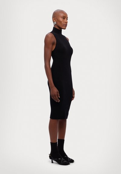 Rochie fără mâneci, din tricot texturat negru, cu un decolteu înalt și o siluetă ajustată, care ajunge până la jumătatea gambei. Este asortată cu șosete negre și pantofi cu toc cu barete.