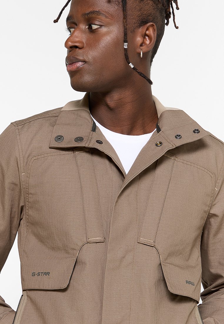 Chaqueta ligera marrón con un diseño estructurado, que presenta un cuello alto, botones a presión y dos bolsillos frontales. Tejido texturizado con costuras visibles.