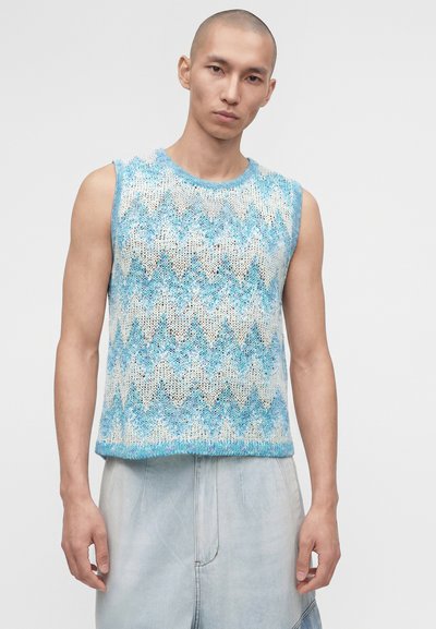 Άνδρας με μπλε και άσπρο πλεκτό τοπ χωρίς μανίκια με ζιγκ-ζαγκ σχέδιο και ανοιχτό μπλε παντελόνι με φαρδιά μπατζάκια, στέκεται σε απλό φόντο.