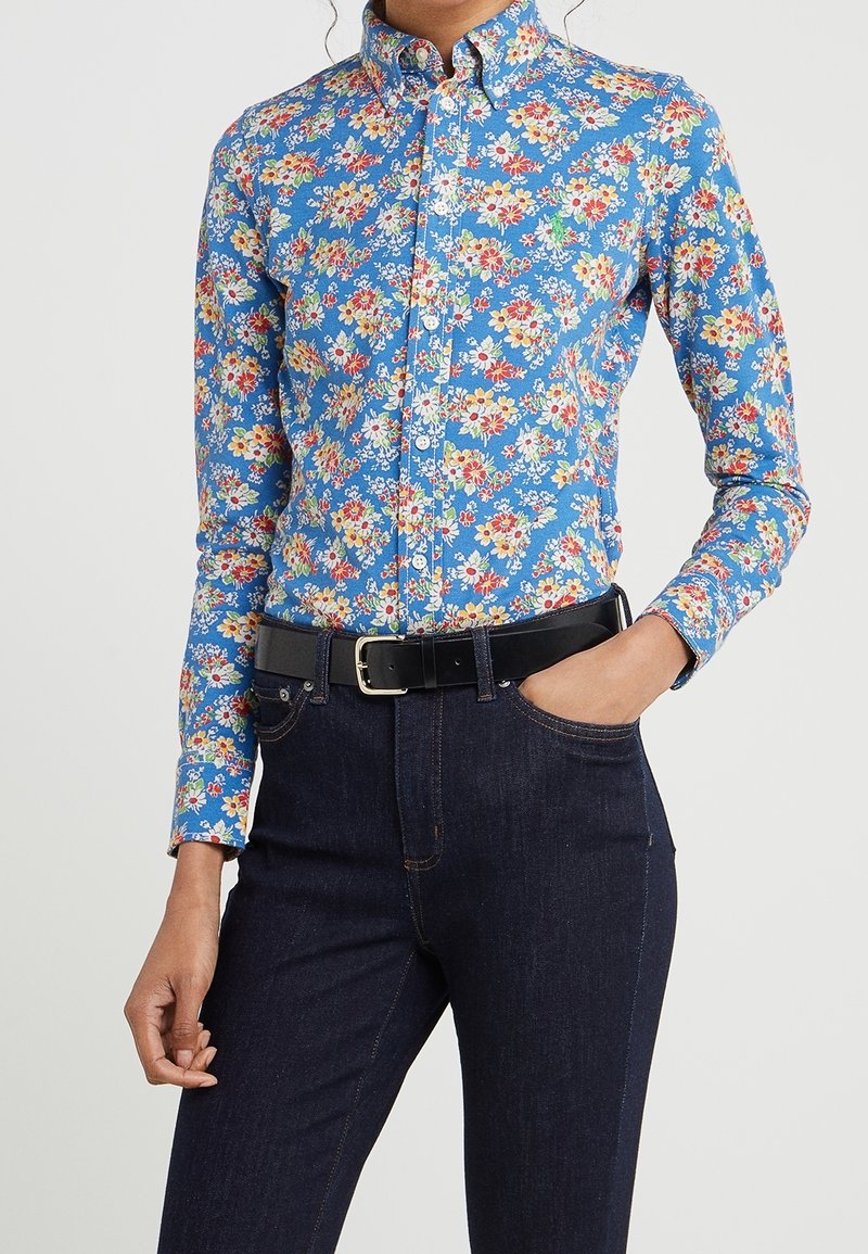 Femme portant une chemise bleue cintrée à boutons avec un imprimé floral multicolore, rentrée dans un jean bleu foncé avec une ceinture noire.