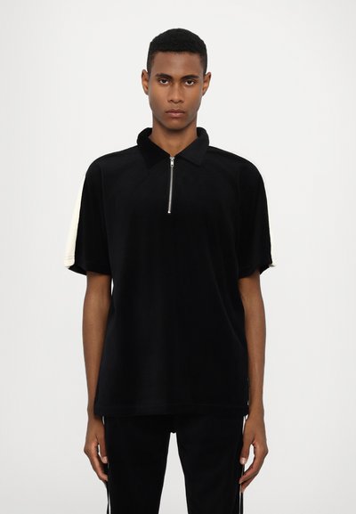 Polo-shirt noir à fermeture éclair, en tissu doux et texturé, avec des manches courtes et des panneaux crème contrastants sur les épaules.