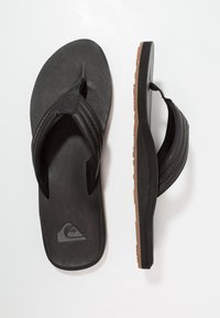 Svarte flip-flopper med teksturert læroverdel, to stropper og en brun gummisåle med geometrisk mønster. Logo på innersålen.