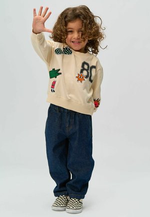 Sudadera beige con parches coloridos, incluyendo un cocodrilo y la impresión "80", combinada con pantalones vaqueros anchos de color azul oscuro y zapatos de lona de cuadros.