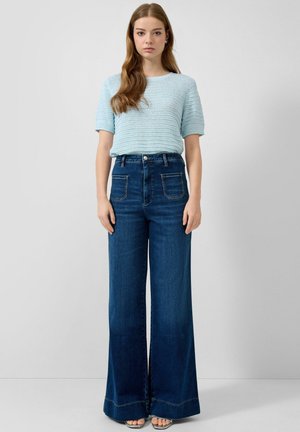 Młoda kobieta z długimi włosami, ubrana w jasnoniebieski krótki sweter z dzianiny i szerokie, wysokociśnieniowe, ciemnoniebieskie dżinsy, stojąca na jednolitym tle.