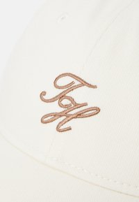 Kremfarget caps med buet brem; har en snekrift brun brodert logo foran; teksturert stoff med synlig søm.